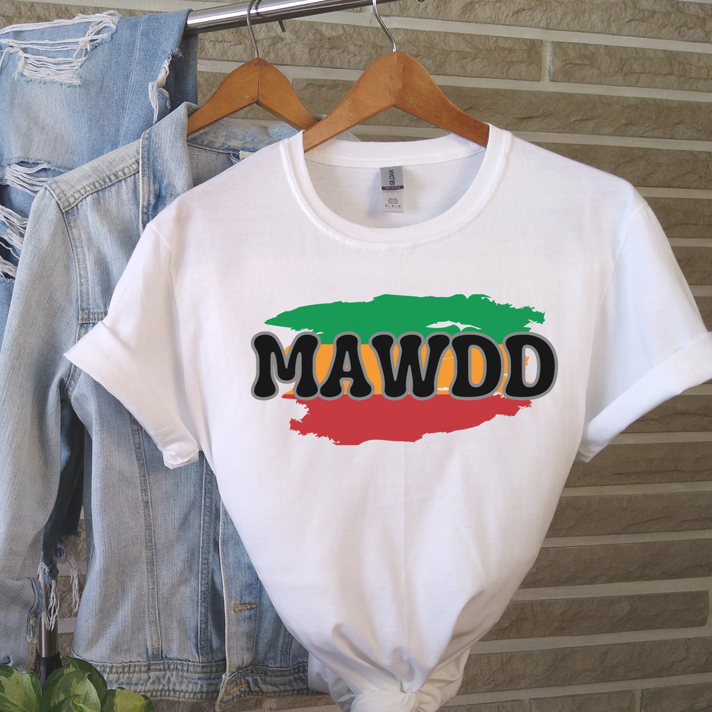 White t-shirt with 'MAWDD' design on a hanger against a brick wall.