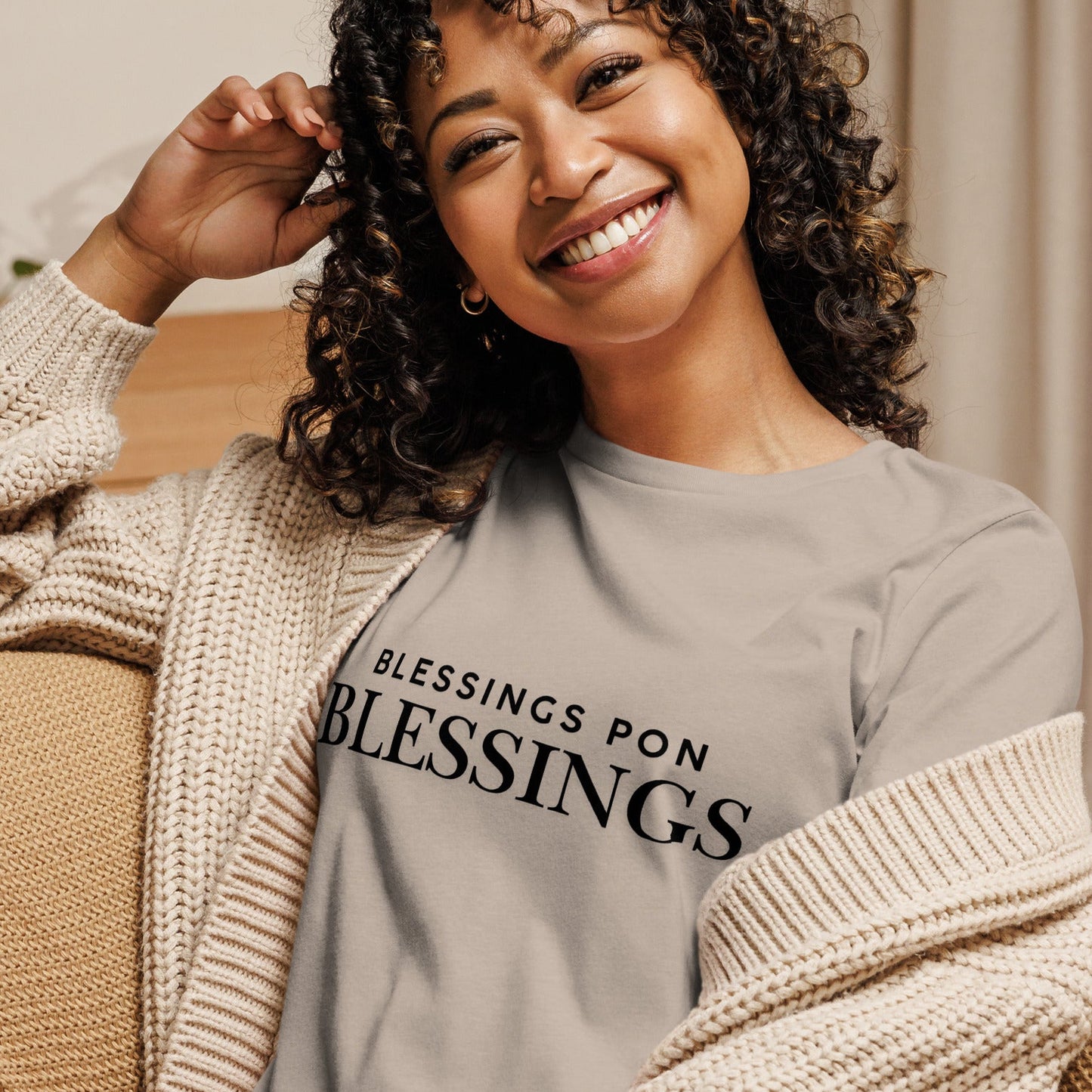 Woman wearing a heather stone t-shirt with 'BLESSINGS PON BLESSINGS' text, sitting on a couch.