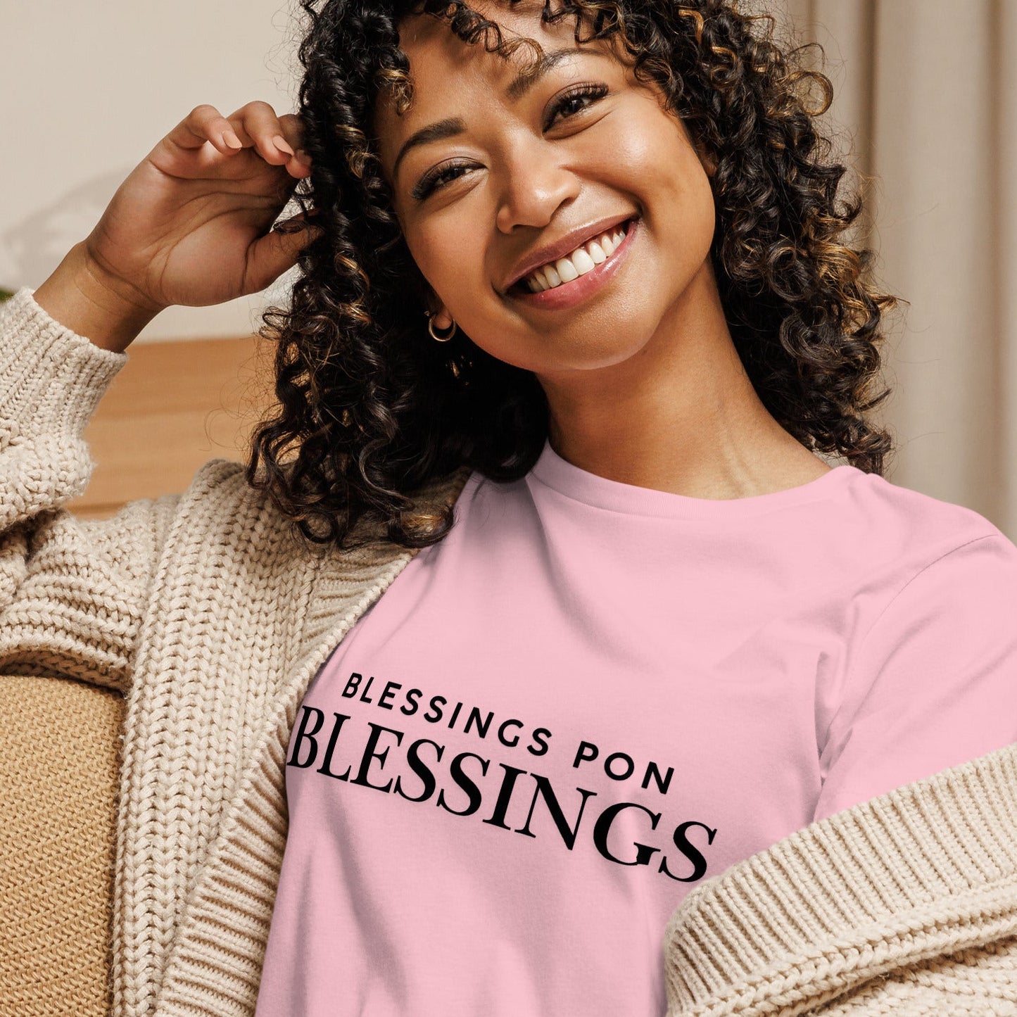 Woman wearing a pink sweatshirt with 'BLESSINGS PON BLESSINGS' text, sitting on a couch.